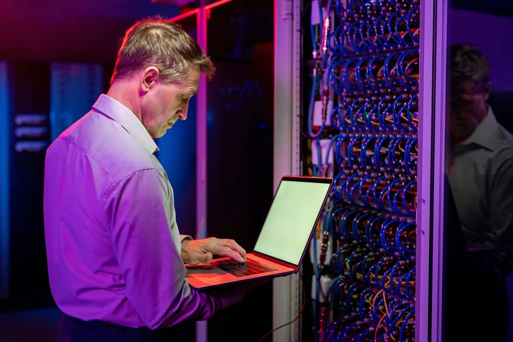 A man in a shirt using a laptop in a server room assessing the poor workplace wifi connection.